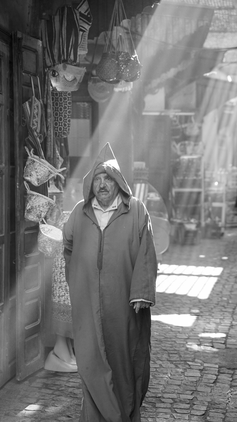 A black-and-white portrait of a man in a hooded robe on a market street with shafts of sunlight